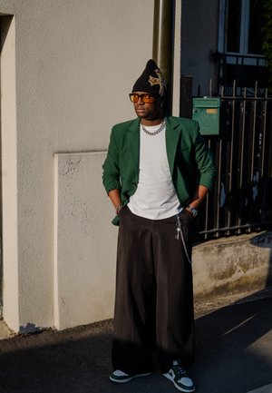Blazer verde, camiseta blanca, gorro negro, pantalones anchos negros, cadena plateada, zapatillas verdes y blancas. Entorno urbano con colores de fondo apagados.