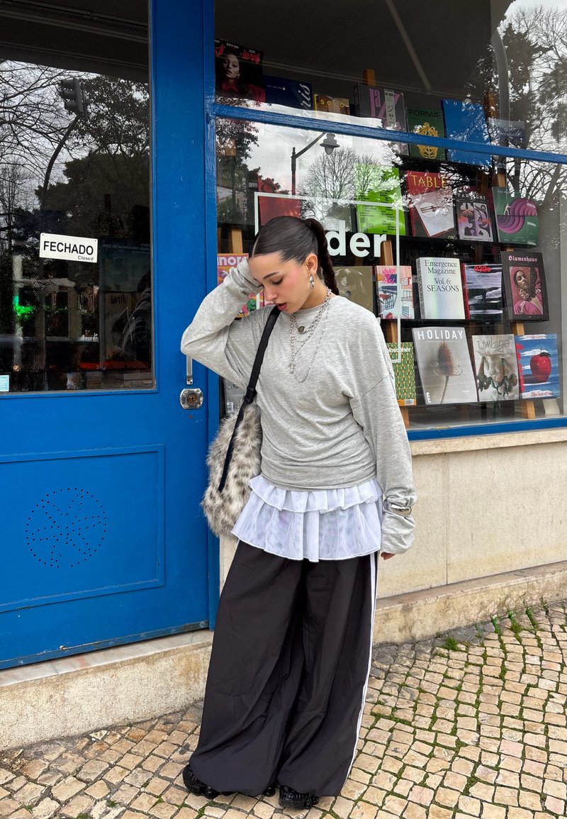 Jeune femme aux cheveux noirs attachés en queue de cheval, portant un pull gris, une jupe blanche superposée et un large pantalon noir, se tient devant une librairie avec une porte bleue.