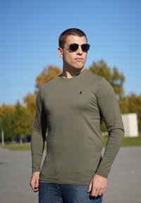 Maglietta verde oliva a maniche lunghe realizzata in tessuto morbido, design aderente, scollo rotondo e piccolo logo. Indossata con jeans blu e occhiali da sole.