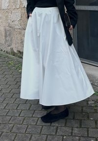A person standing on a brick pavement, wearing a long white skirt, a black jacket, black flats, and holding a small black handbag.