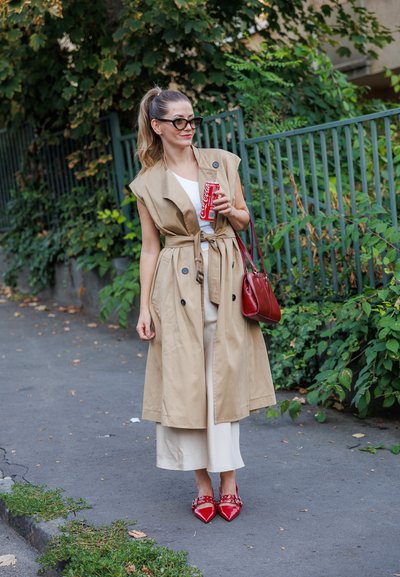 Gabardina sin mangas beige con cinturón, top blanco, pantalones crema, bolso rojo y zapatos puntiagudos rojos con tachuelas. Sosteniendo una lata de refresco.