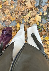 Veste en cuir marron, pantalon large blanc, bottines grises, et un sac à main tissé bordeaux, le tout sur un fond de feuilles d'automne tombées.