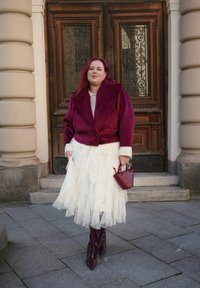 Burgundi hamis szarvasbőr kabát széles gallérral, réteges fehér tüll szoknya, burgundi vödörtáska és hegyes orrú, térdig érő csizma.