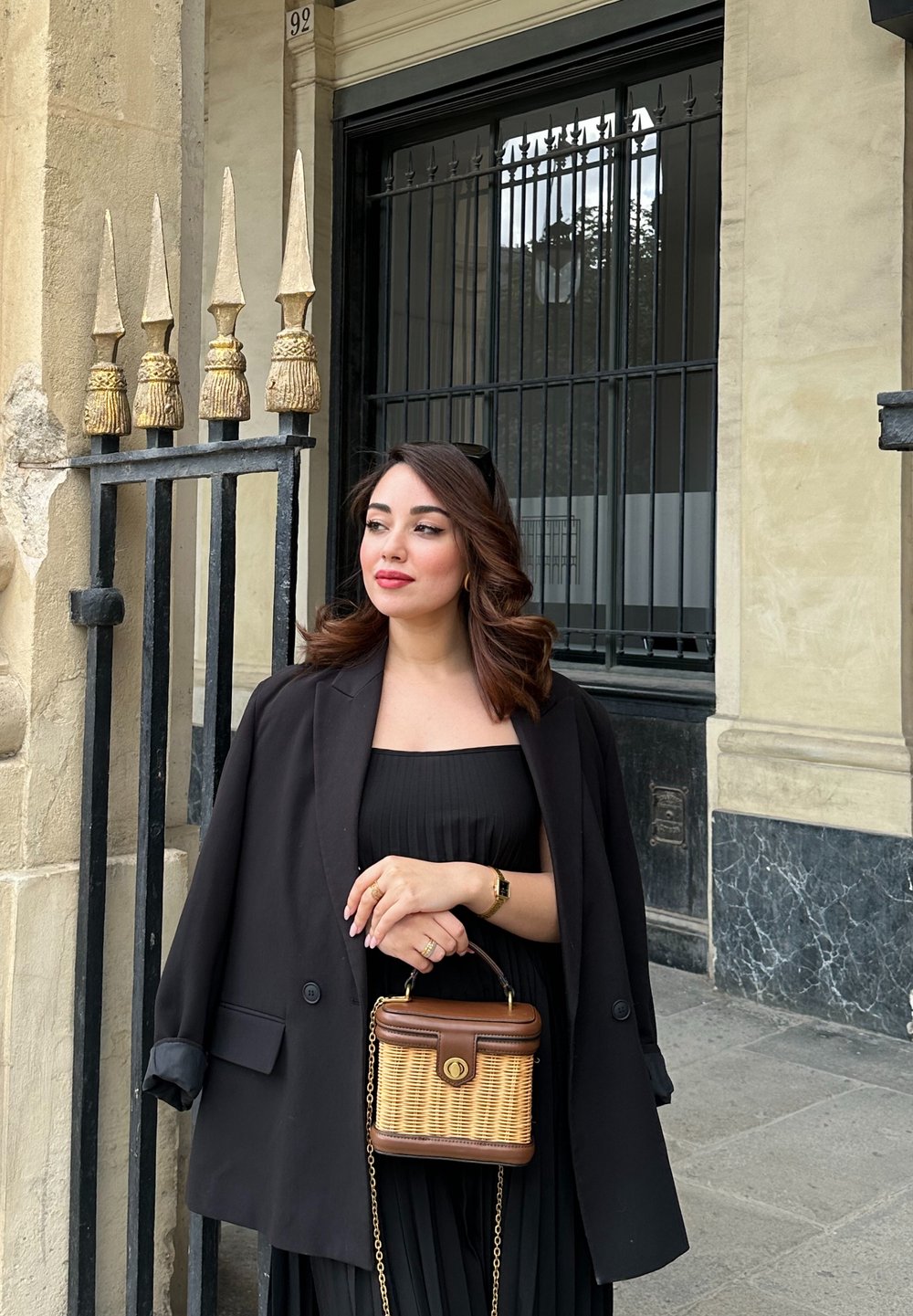 Schwarze übergroße Blazer, plissiertes schwarzes Kleid und eine braune Korbtasche mit goldenen Hardware-Akzenten. Dekorativer Zaun im Hintergrund.