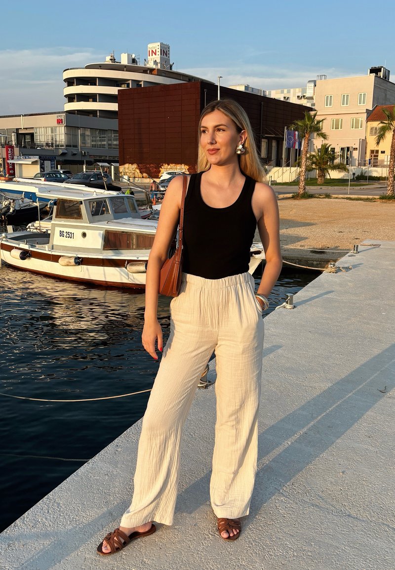 Débardeur noir à côtes associé à un pantalon ample plissé beige clair et des sandales marron. Petit sac à main marron et cadre naturel au bord de l'eau.