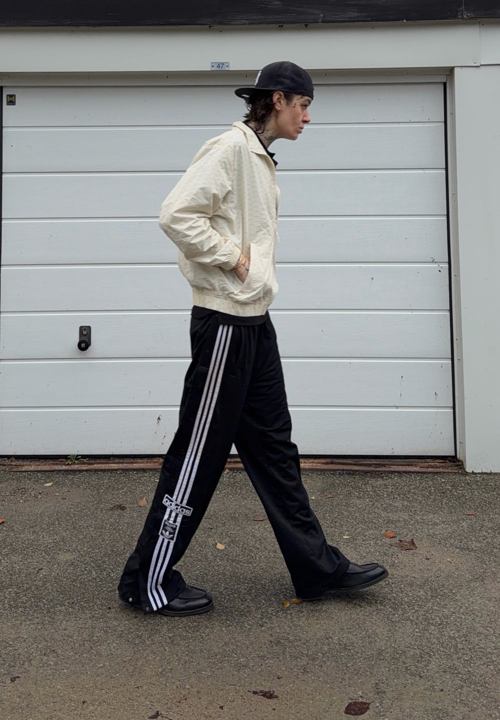 Light beige jacket with zip closure, black sports pants with white side stripes, and black trainers. Man walking in front of a garage door.