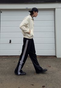 Light beige jacket with zip closure, black sports pants with white side stripes, and black trainers. Man walking in front of a garage door.