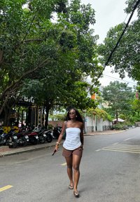 Top blanco sin tirantes, pantalones cortos marrones y chanclas. Cabello largo y oscuro, gafas de sol, sosteniendo un teléfono, caminando por una calle bordeada de árboles.