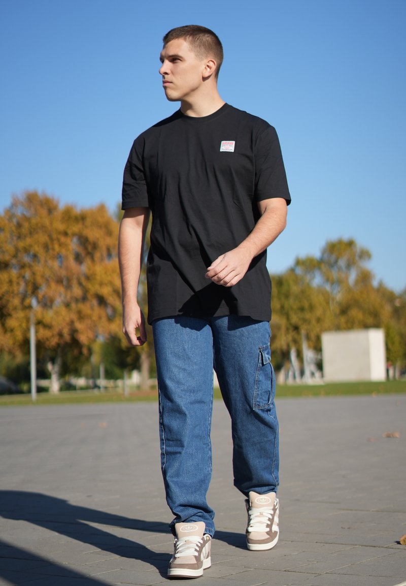 Sort bomulds T-shirt med korte ærmer og et logo-patch, parret med blå cargo-jeans med sidelommer og lyse sko.