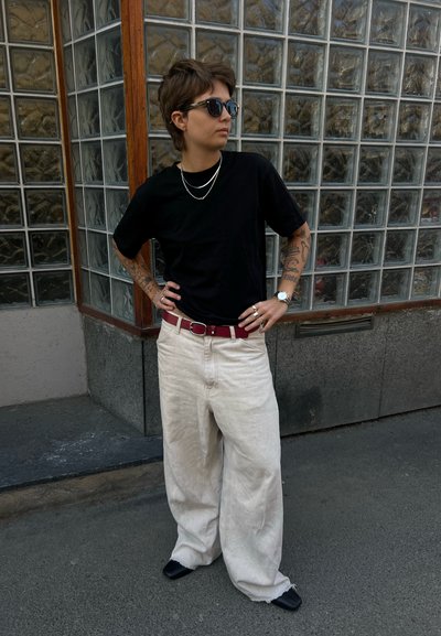 Persona con el pelo corto, que lleva gafas de sol, camisa negra, pantalones anchos beige, cinturón rojo y tatuajes en los brazos, de pie con las manos en las caderas.