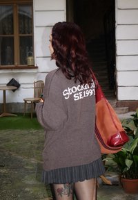 Brun oversized sweater med teksten "Stockholm SE 1991" på ryggen, parret med en sort plisseret nederdel og en rødbrun taske.