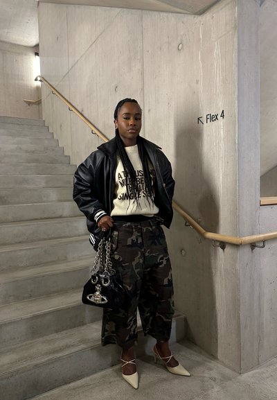 Mujer con trenzas que lleva una chaqueta de cuero negra, un suéter blanco, pantalones de camuflaje y tacones blancos, de pie junto a escaleras de concreto y un letrero en la pared que dice "Flex 4."