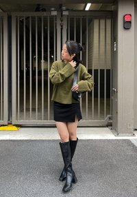 Mulher de suéter verde, mini saia preta e botas pretas até ao joelho, em pé no alcatrão em frente a um portão metálico fechado com uma luz vermelha.