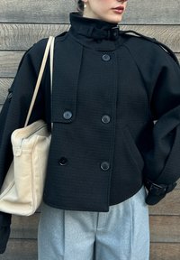 Sort dobbeltradet jakke med høj krave, lavet af tekstureret materiale, med store knapper og taljebælte. Lysfarvet taske.