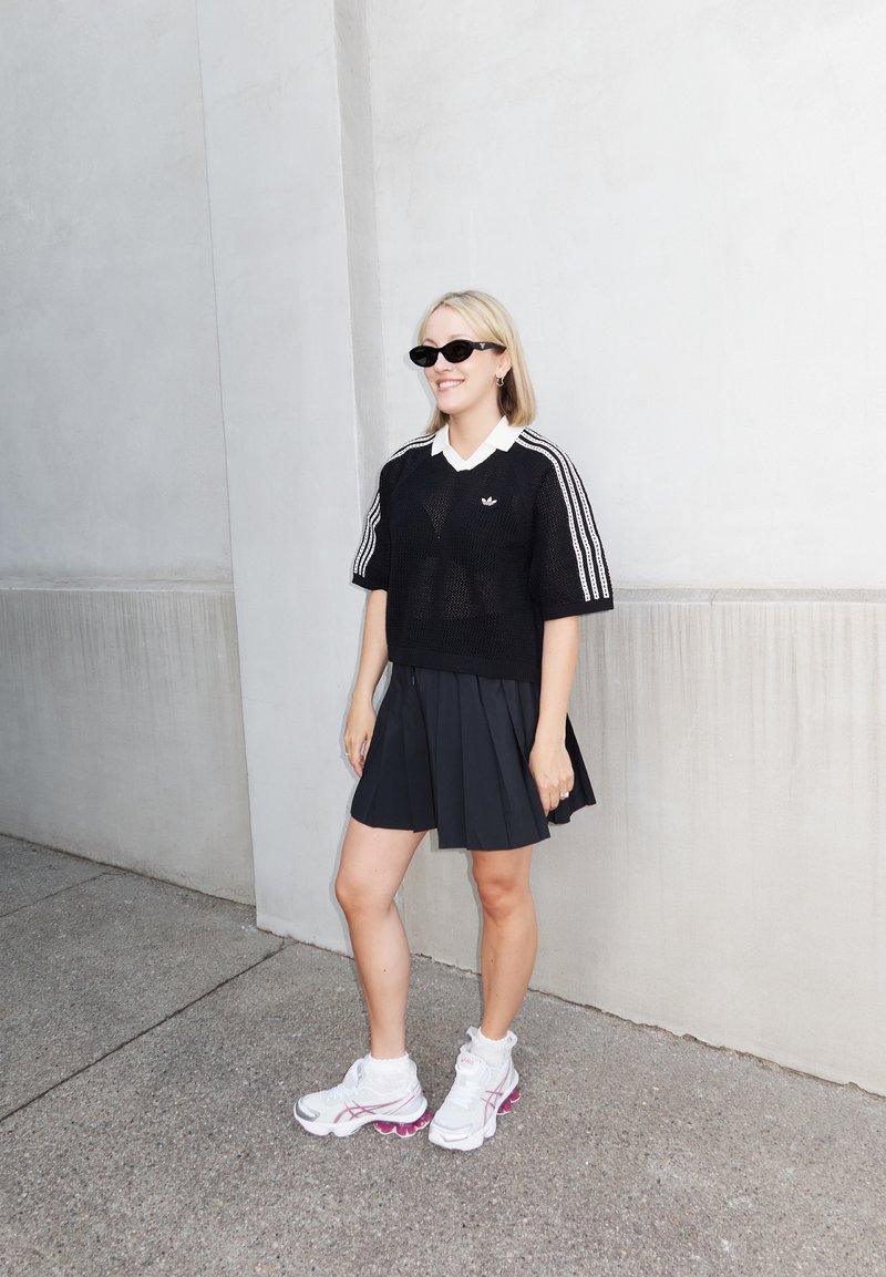 Γυναίκα με μαύρο διαπνέον τοπ Adidas, πλισέ φούστα, λευκά αθλητικά παπούτσια και γυαλιά ηλίου στέκεται δίπλα σε λευκό τοίχο σε τσιμεντένιο πεζοδρόμιο.