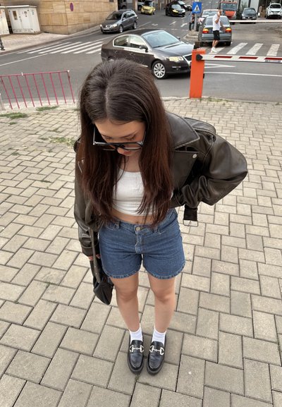 Chaqueta de cuero negro sobre una blusa blanca corta, con pantalones cortos de mezclilla azul. Lleva mocasines negros y calcetines blancos. De pie sobre un pavimento de ladrillos.