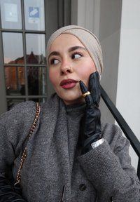 Femme portant un foulard beige, un manteau gris et des gants en cuir noir avec une bague en or, reposant la main sur son visage, debout près d'une porte vitrée avec des reflets.