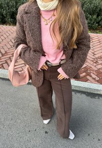 Blazer marron et duveteux sur un pull rose, associé à un pantalon marron et des chaussettes blanches à motifs. Tient un sac rose texturé. Collier en or avec des charms.