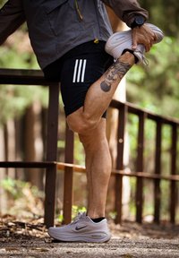 Mees, kes kannab musti lühikesi pükse ja valgeid spordijalanõusid, venitades oma tätoveeritud jalga, hoides jalast kinni selja taga metsasteel.