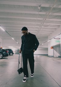 Črn suit in hlače v kombinaciji s črnim turtleneckom, nosi majhno črno torbico. Črne teniske z belim podplatom. Notranji parkirni prostor.