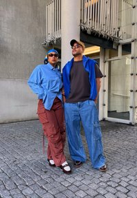 Due persone sono in piedi su una superficie di ciottoli. Uno indossa una camicia azzurra con bottoni, pantaloni cargo marroni e scarpe bianche. L'altro indossa un gilet blu sopra una maglietta nera e pantaloni di jeans larghi. Entrambi indossano occhiali da sole.