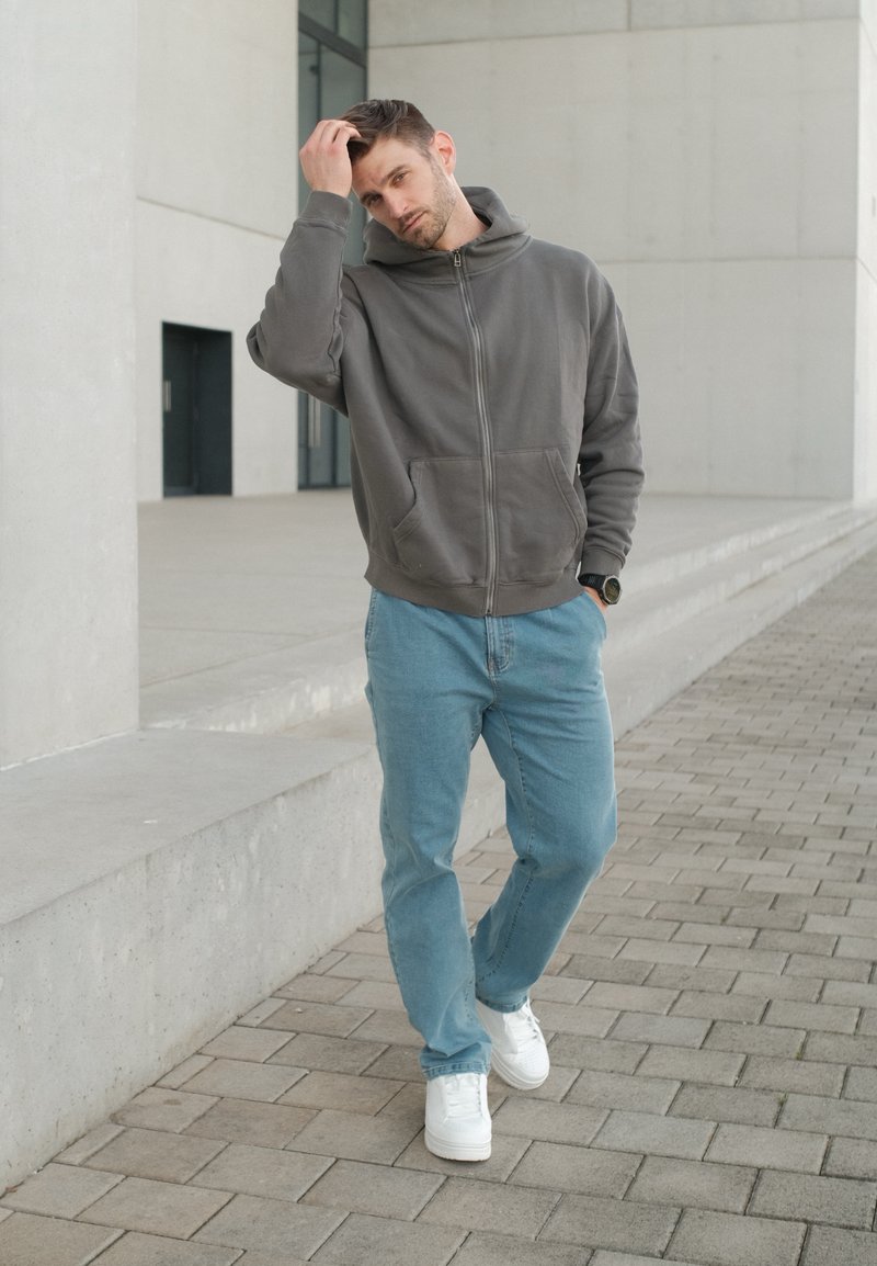 Homme en sweat à capuche gris et jean bleu debout sur un chemin pavé, touchant ses cheveux d’une main près d’un bâtiment moderne en béton.