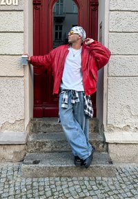 Červená kožená bomber bunda, bílý tričko, volné modré džíny, černobílá kostkovaná košile uvázaná v pase, sluneční brýle, bandana, drží hrnek s kávou.