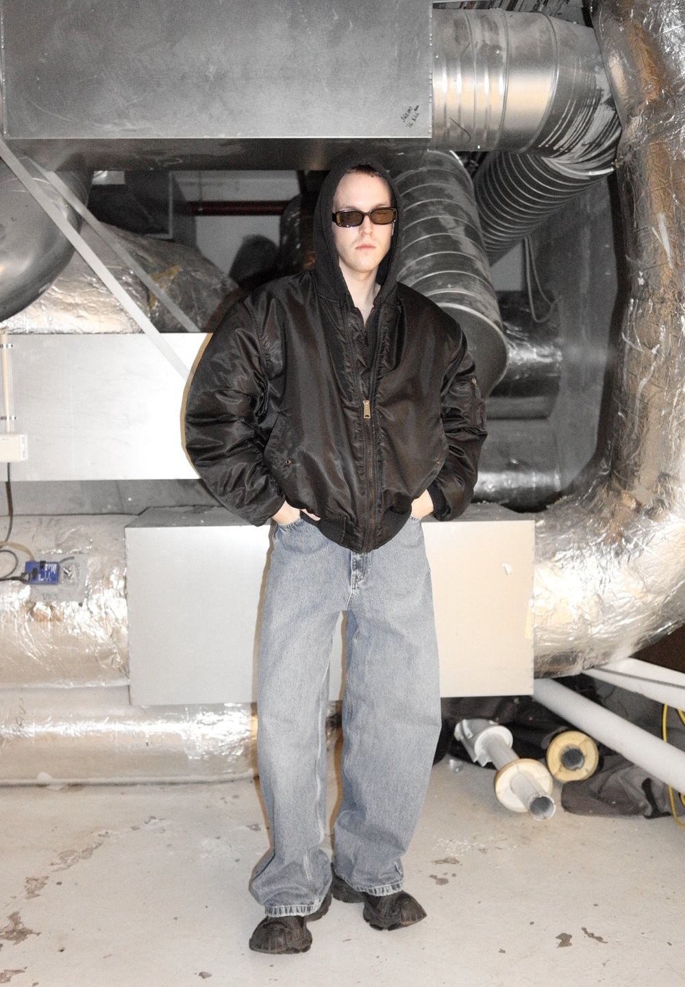 Giacca bomber nera con zip, jeans larghi azzurri, scarpe nere e occhiali da sole. Lo sfondo presenta condotti metallici e superfici grigie.