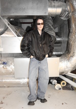 Chaqueta bomber negra con cremallera, vaqueros holgados azul claro, zapatos negros y gafas de sol. El fondo presenta conductos metálicos y superficies grises.