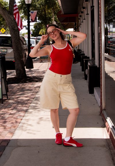 Top deportivo sin mangas rojo con ribete blanco, pantalones cortos beige y sneakers rojos. Las gafas y un bolso negro completan el look. Ambientación al aire libre.