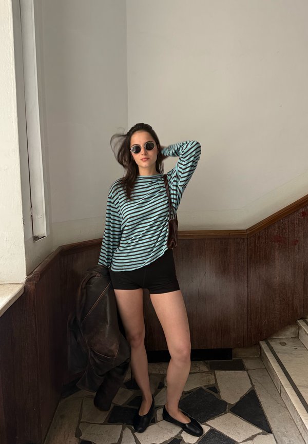 Femme portant des lunettes de soleil, un chemisier rayé à manches longues et un short noir, debout à l'intérieur tenant une veste à ses côtés.