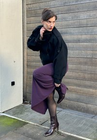 Veste noire oversize, jupe asymétrique violette, collants noirs transparents et bottes à talons marron. Le mannequin pose avec la main sur le col devant un fond en bois.