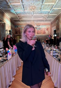 Jeune femme en blazer noir sirotant un cocktail dans une pièce ornée avec de longues tables de banquet, des lustres et des fresques murales, entourée d'invités.