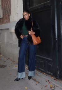 Mustikka keinoturkki takki, vihreä neule, leveälahkeiset farkut, joissa on erottuvat lahkeet, mustat teräväkärkiset kengät ja oranssi käsilaukku tummaa seinää vasten.