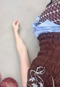 Haine dintr-un material texturat de culoare maro, cu broderie florala; legături de accent de culoare bleu deschis; un braț întins cu piele moale, așezat pe o suprafață gri.