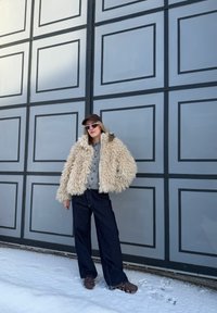 Personne portant une veste beige duveteuse, un cardigan gris, un pantalon large foncé, des chaussures marron, des lunettes de soleil roses et une casquette marron, debout sur la neige devant un mur gris à panneaux.