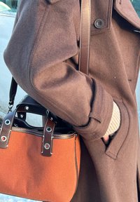 Personne portant un manteau en laine marron avec la main dans la poche, portant un sac à main en tissu orange structuré avec des anses en cuir marron foncé.