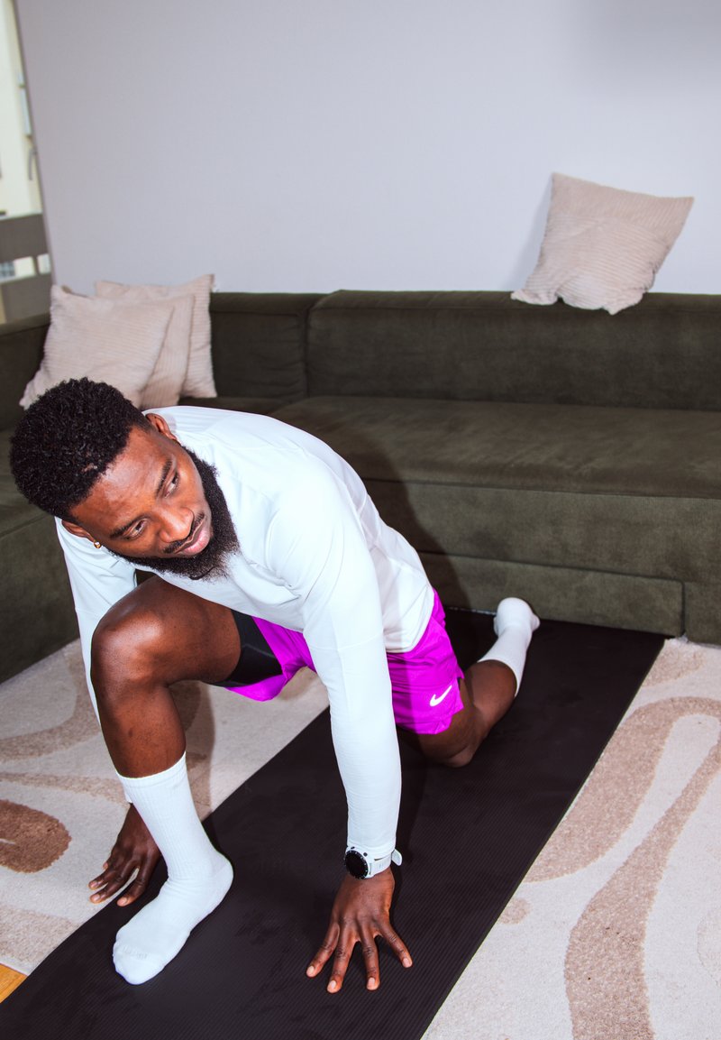 Homme en chemise blanche à manches longues et short rose s'étirant sur un tapis noir dans un salon avec un canapé vert foncé et des coussins beiges.