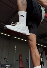 Weißer Sportschuh mit strukturierter Oberfläche und rosa gefärbter Sohle, kombiniert mit einer weißen Socke mit schwarzem Logo. Kurze schwarze Shorts sind sichtbar.