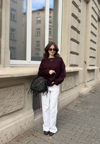 Bordo džemper s otvorenim ramenima, bijele široke hlače, crna torba s resama, tamne cipele, stoji pored teksturiranog zida i prozora.