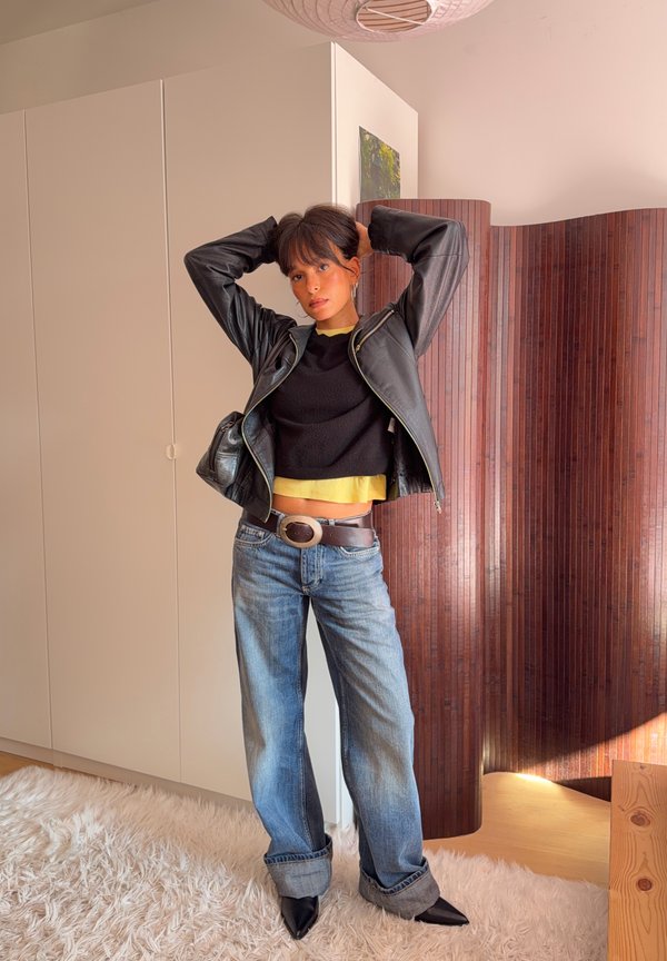 Una mujer posa con las manos detrás de la cabeza. Lleva una chaqueta de cuero negra, un top amarillo, vaqueros con el bajo vuelto, un cinturón y zapatos de punta.