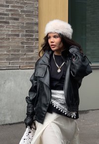 Schwarze, übergroße Lederjacke, weißer Pelzhut, silberne Halskette, schwarzes Oberteil, weißer Rock mit Spitzenakzenten, schwarze Handschuhe, gemusterte Tasche.