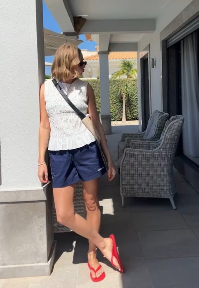 Top sin mangas texturizado blanco, pantalones cortos azul marino, chanclas rojas; de pie junto a pilares blancos con sillas de mimbre en un entorno exterior soleado.