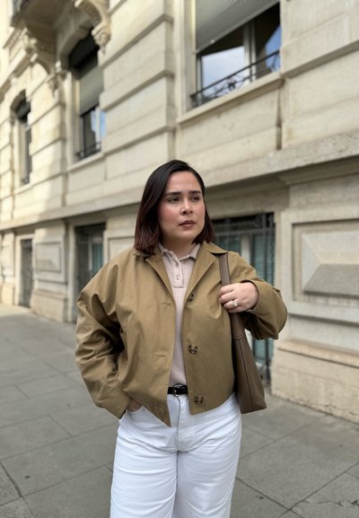 Chaqueta corta marrón claro sobre un polo beige, pantalones blancos de cintura alta, sosteniendo un bolso marrón. Texturas suaves, detalles de botones sutiles.