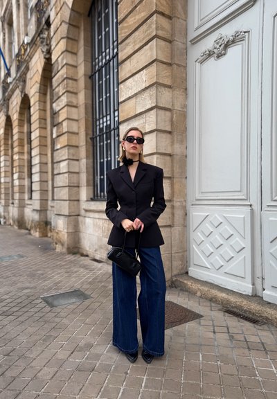 Blazer negro entallado con jeans de pierna ancha de mezclilla. Los accesorios incluyen un bolso negro y gafas de sol, todo contra una arquitectura de piedra.