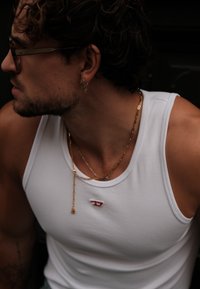 Top de tirantes de algodón blanco con un pequeño logotipo rojo, cadenas de oro en capas y pendientes de aro plateados. Se pueden ver cabellos rizados y piel texturizada.