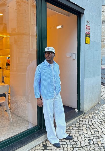 Hombre con camisa de botones a rayas azules y blancas, pantalones blancos y gorra gris, de pie en una puerta abierta con suelo de baldosas.