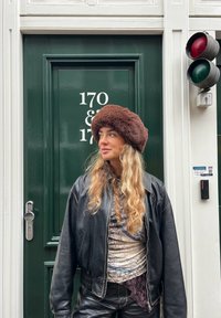 Femme aux longs cheveux blonds portant un chapeau en fourrure marron et une veste en cuir noire, debout devant une porte vert foncé numérotée 170 & 172.