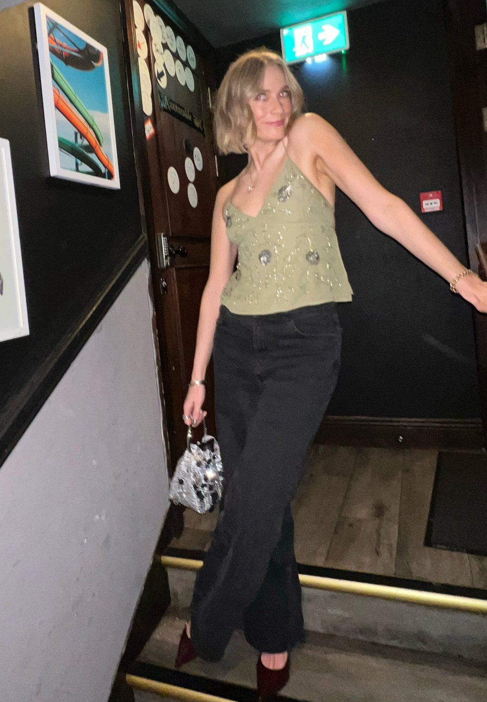 Haut sans manches vert orné de sequins, pantalon large noir, et sac à main argenté brillant, porté avec des talons bordeaux, sur un fond faiblement éclairé.