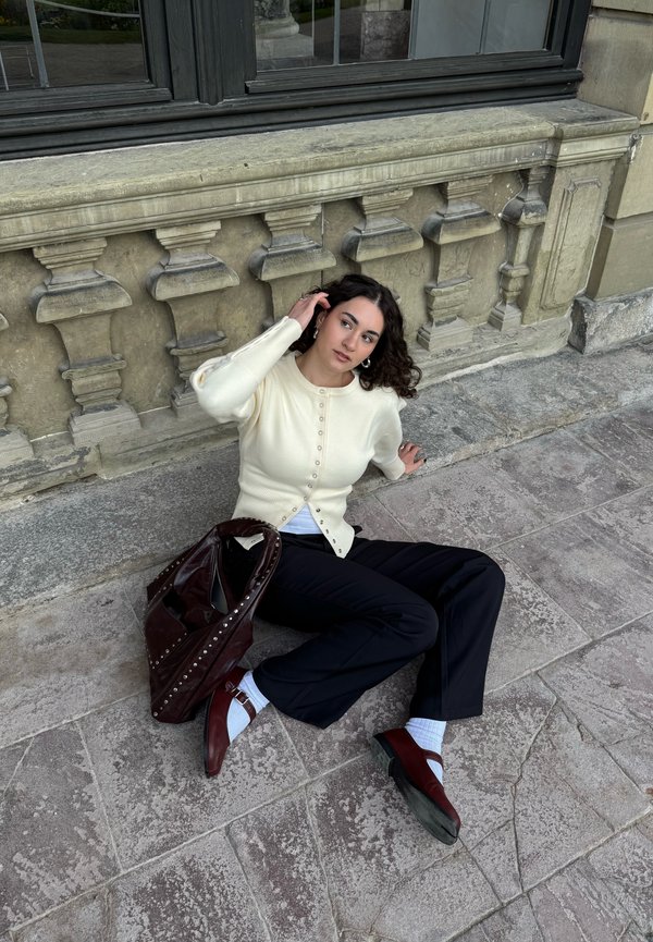 Femme aux cheveux bouclés et foncés assise sur un pavé en pierre, appuyée contre un mur de pierre orné, portant un pull crème, un pantalon noir et des chaussures bordeaux.